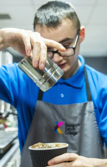 A young barista in a blue uniform sprinkles cocoa powder over a freshly prepared cup of coffee in a café setting. A young barista in a blue uniform sprinkles cocoa powder over a freshly prepared cup of coffee in a café setting.